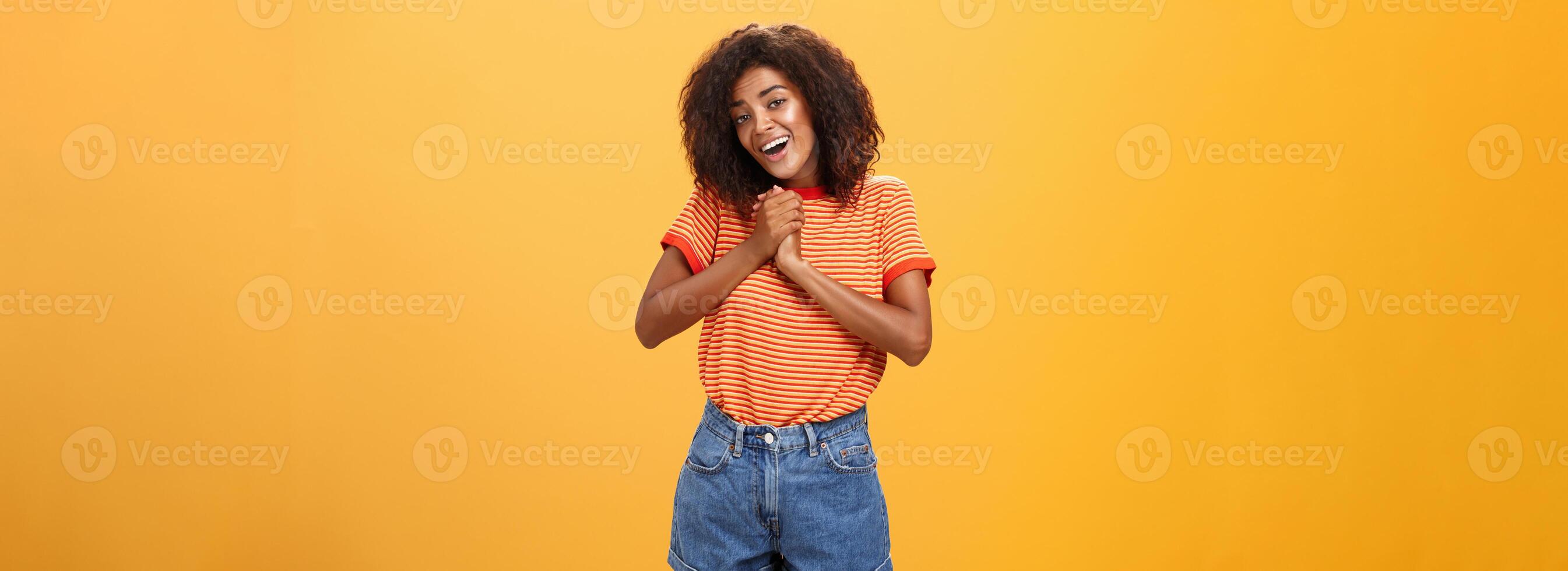 niña suspirando y mirando a linda cosa apretando palmas juntos cerca corazón sonriente con admiración y amor siendo femenino y oferta posando terminado naranja antecedentes sensación afecto foto