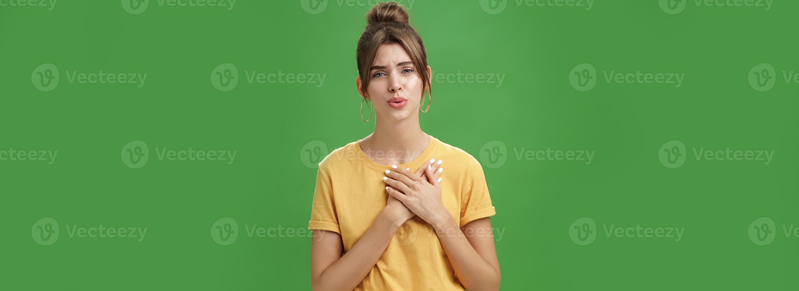 Touched and pleased attractive young woman in yellow t-shirt holding palms pressed to chest in thankful and grateful gesture folding lips melting heart seeing cute thing over green background photo