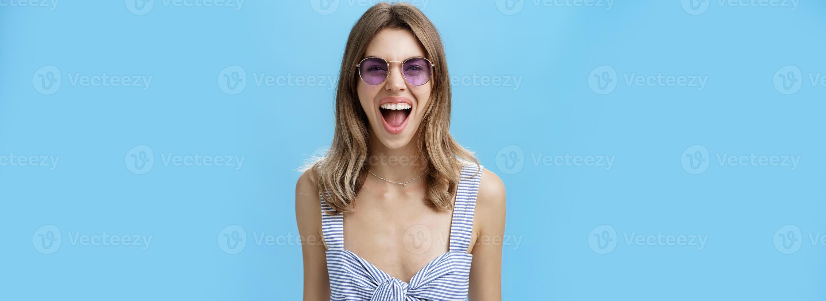 Expressive and careree woman being wild ad free yelling out loud at camera wearing awesome stylish sunglasses and striped top letting out emotions being open-minded and rebellious over blue wall photo