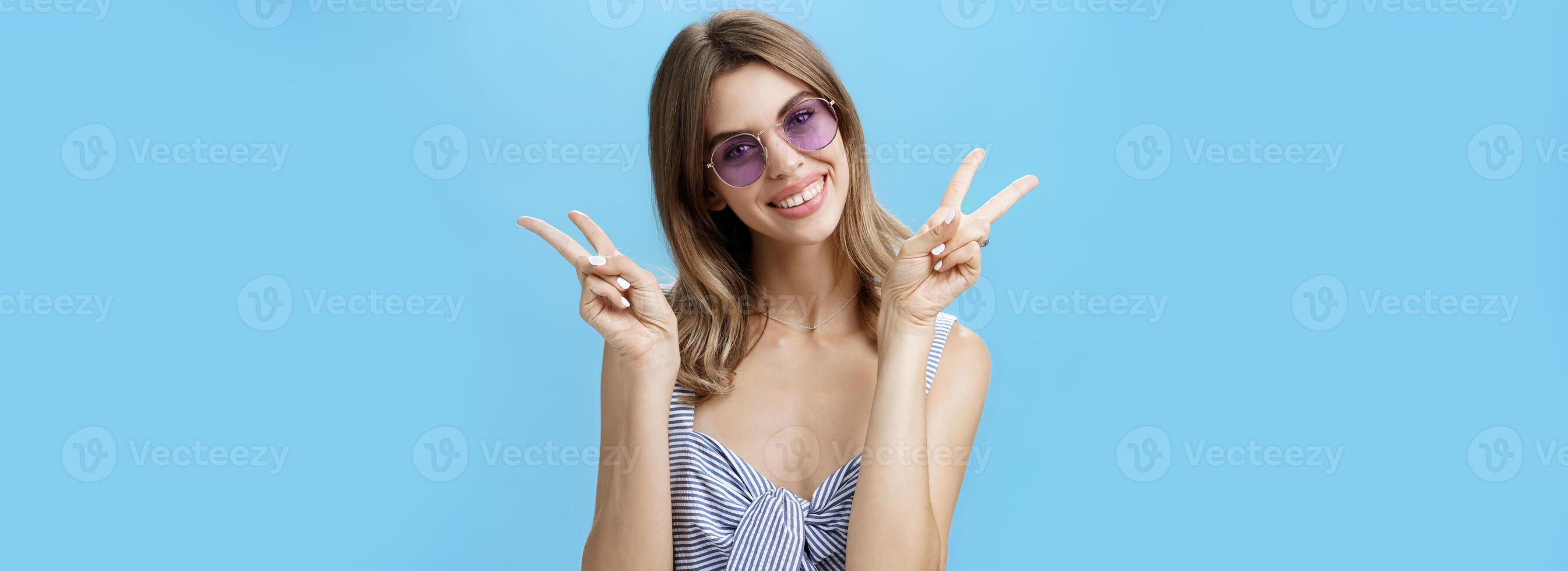 Waist-up shot of cute self-assured feminine girlfriend in trendy top showing peace gestures near shoulders tilting head amused smiling broadly showing off gapped teeth posing over blue wall photo