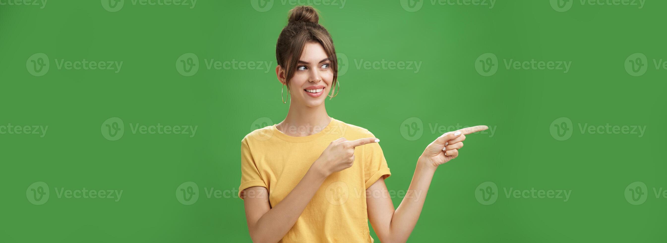 Waist-up shot of good-looking friendly and feminine female coworker in yellow t-shirt showing newbie office pointing and looking left with cute happy smile posing against green background photo