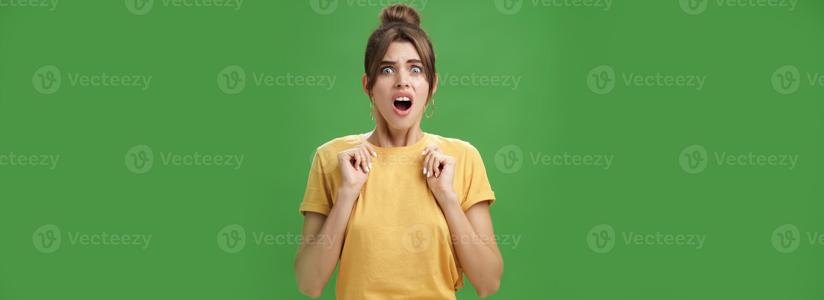 Shocked feminine glamour woman with combed hair in yellow t-shirt frowning gasping with opened mouth holding palms pressed to chest from aversion and disgust looking at creepy thing over green wall photo