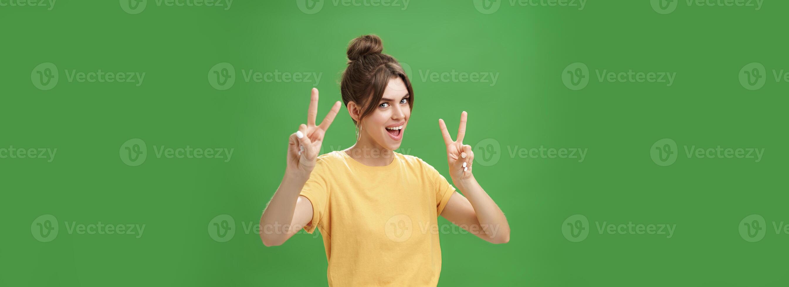 Indoor shot of enthusiastic excited and happy daring girl with combed hair tattoo and cute diasdema showing peace signs bending backwards standing in cool energized pose over green background photo