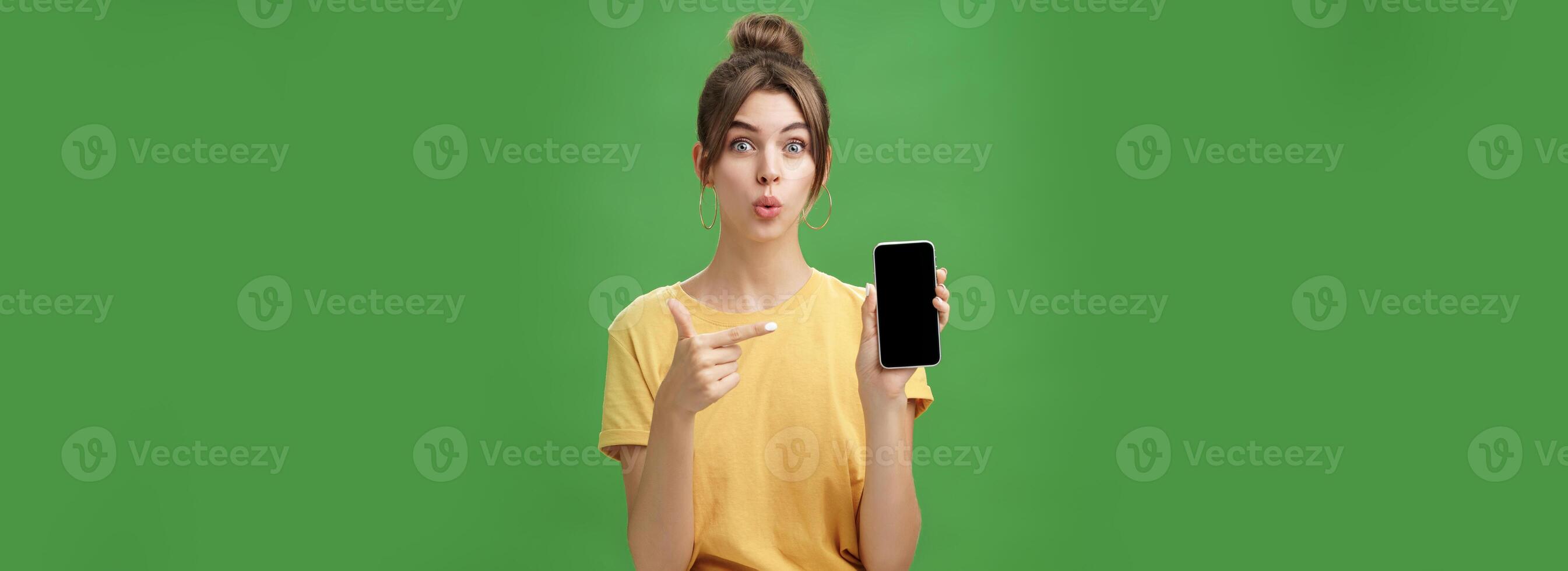 Portrait of intrigued woman cannot wait use new smartphone holding phone and pointing at cellphone screen folding lips with excitement and interest presenting new device model in store over green wall photo