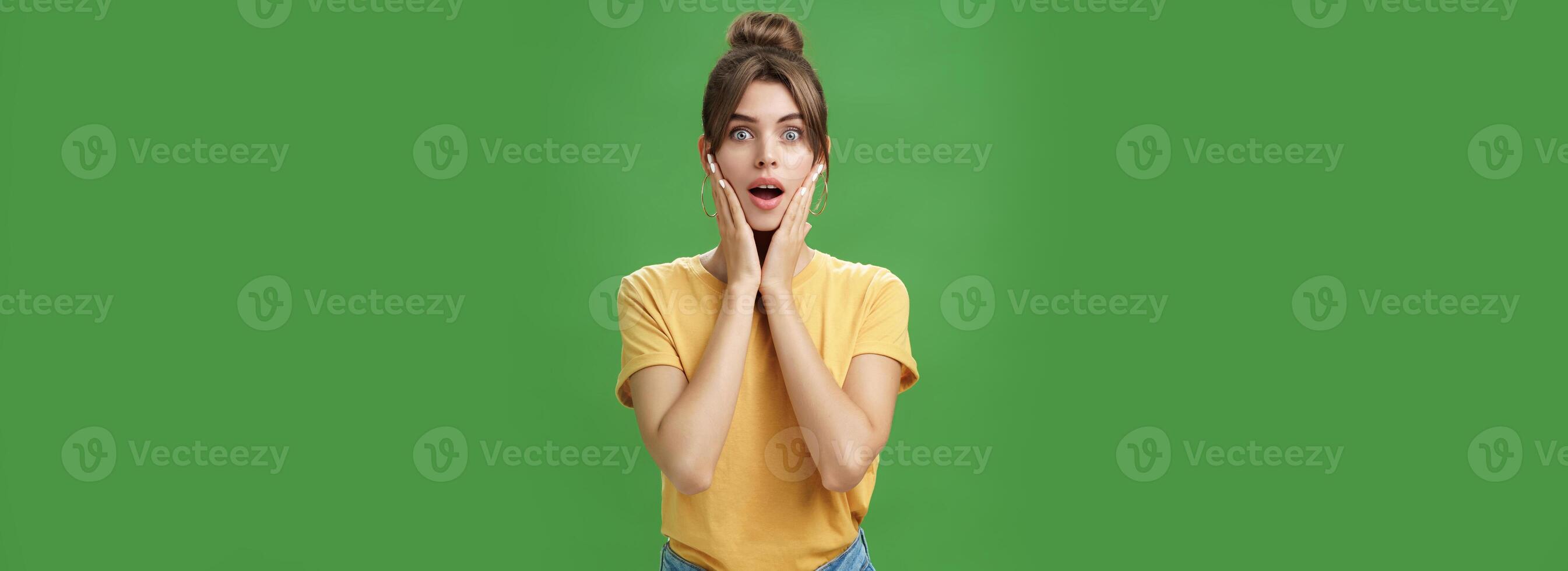 Woman feeling surprised and delighted enjoying good result after detox and applying facial skincare mask touching cheeks opening mouth from amazement and excited posing over green background photo