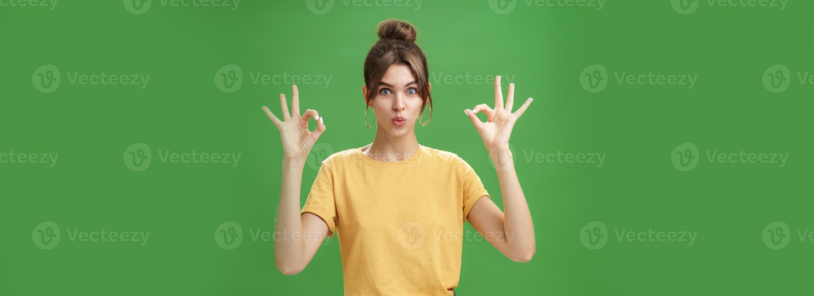 Portrait of silly cute and beautiful caucasian woman with combed hair in round earrings and yellow t-shirt showing okay, no problem gesture folding lips, reaffirming boss job goes well over green wall photo