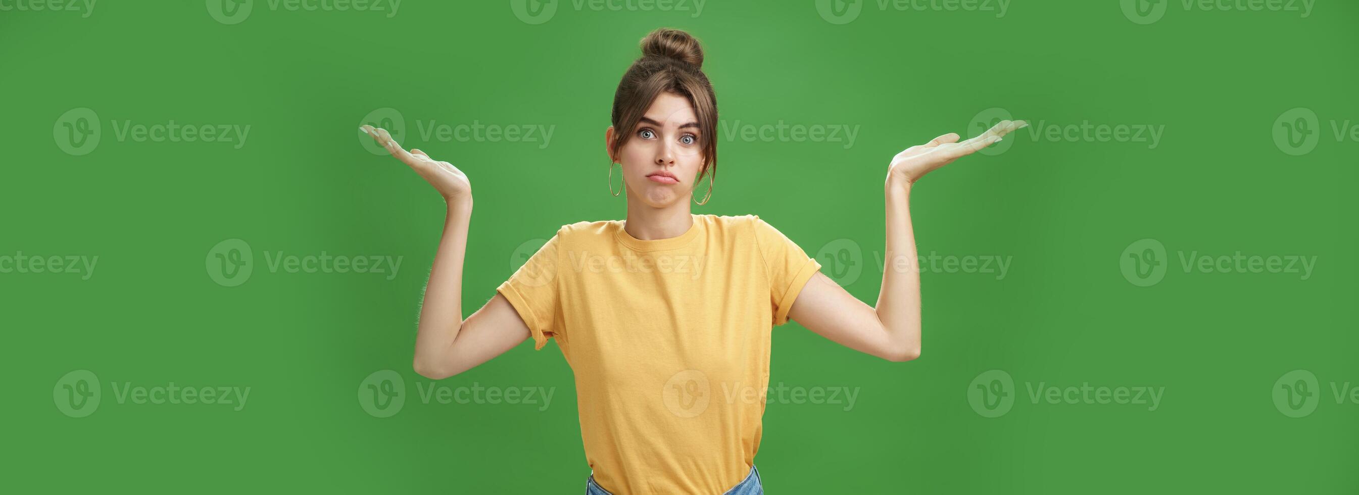 Girl do not know anything. Portrait of confused unsure cute woman with combed hair shrugging with raised hands pouting looking clueless at camera standing questioned against green background photo