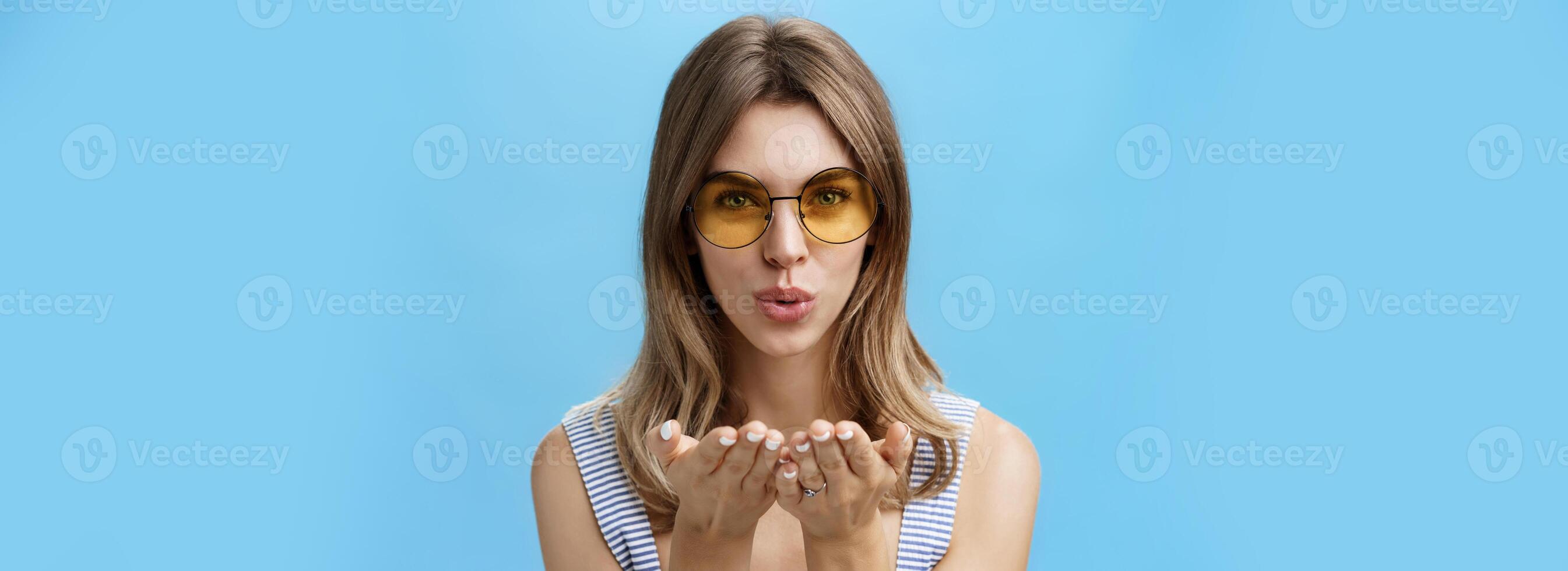 Close-up shot of feminine and tender charming woman in trendy yellow sunglasses folding lips holding palms near mouth to send air kiss at camera gazing sensually forward over blue background photo