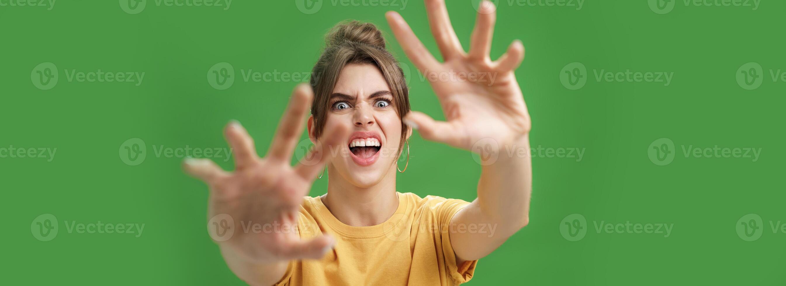 Funny emotive woman making fun face pulling hands forward to attack of grab something frowning opening mouth wanting cover face from camera, grimacing over green background photo