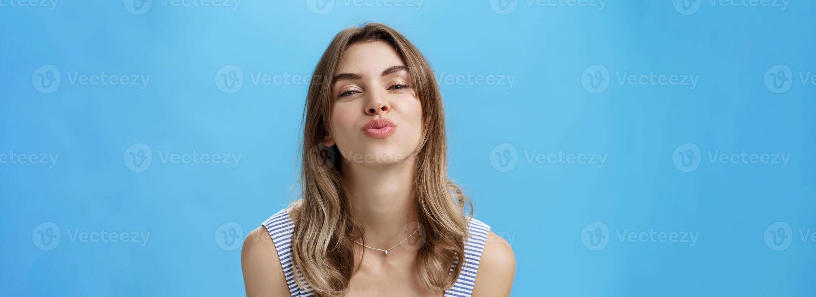 Close-up shot of attractive sensual and flirty young cute woman folding lips in mwah bending towards camera smiling and giving kiss at camera standing happy and self-assured against blue background photo