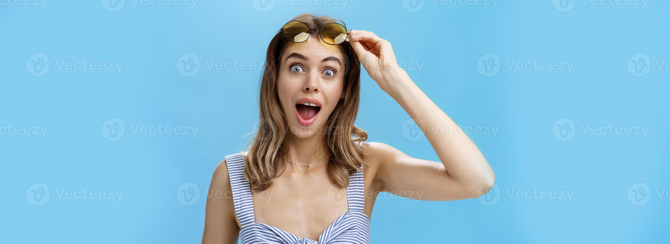 Woman walking across steet meet friend taking sunglasses and gazing surprised at camera opening mouth to say hi with broad happy grin and pleased expression standing amazed over blue wall photo