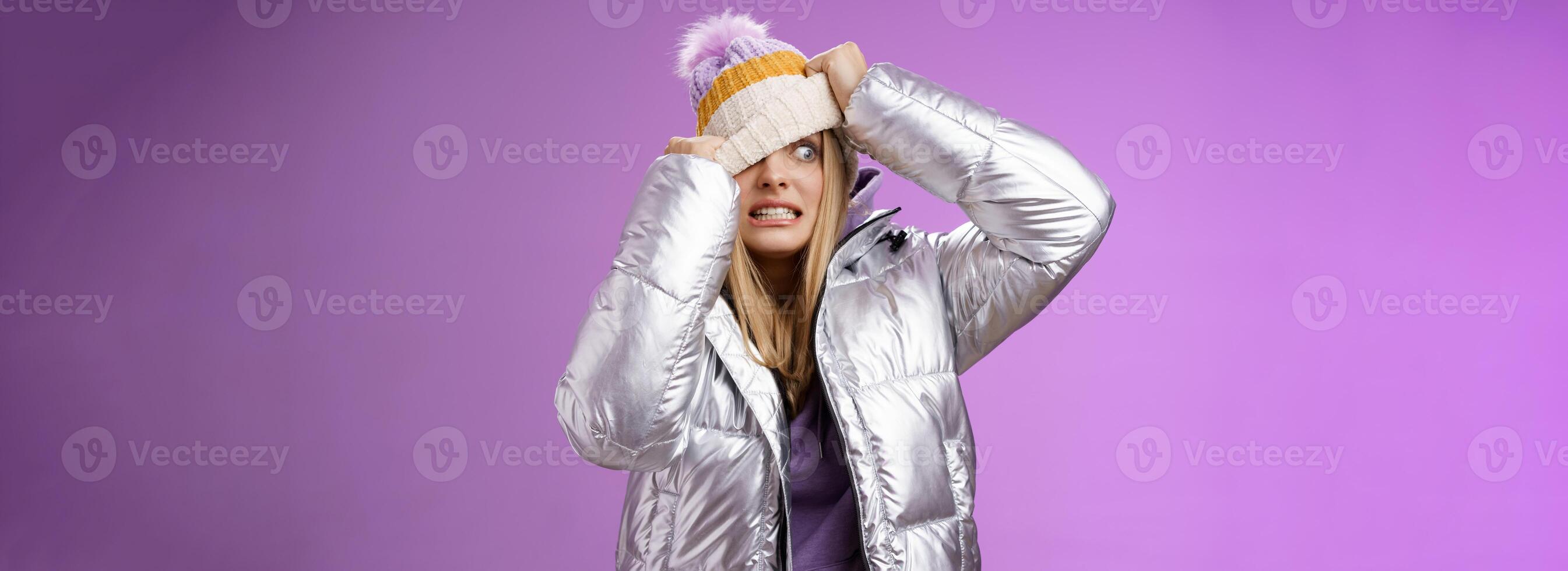 Awkward embarrassed cute blond girl worried pulling hat face hiding from ex-boyfriend pretend not see him standing anxiously clenching teeth peeking one eye left nervous, purple background photo