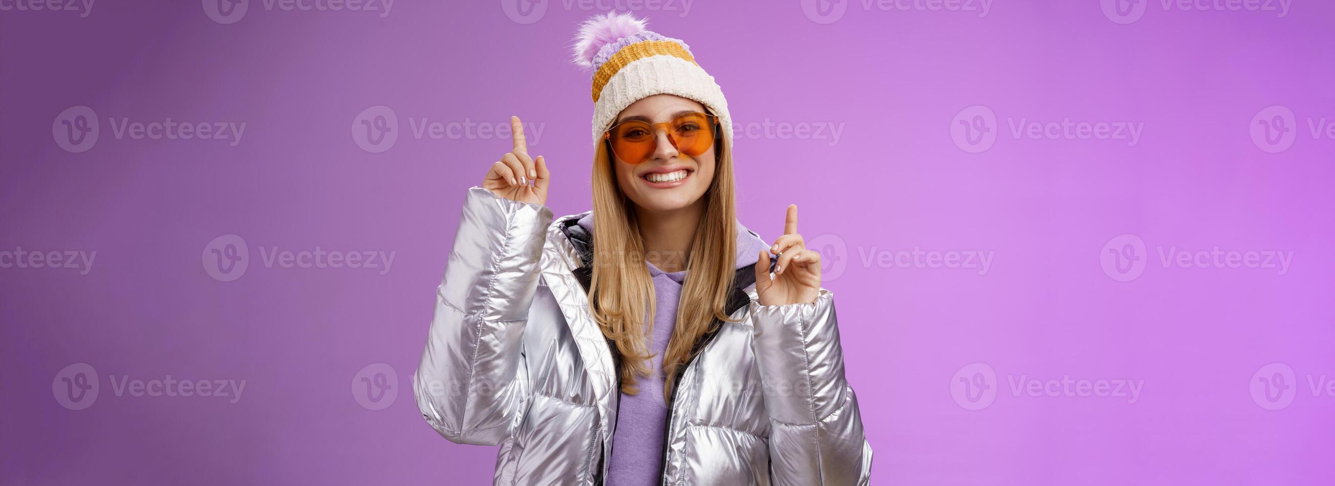 Joyful energized entertained cute blond woman having fun enjoy vacation snowy mountain trip wearing sunglasses silver jacket winter hat dancing pointing up amused standing purple background photo