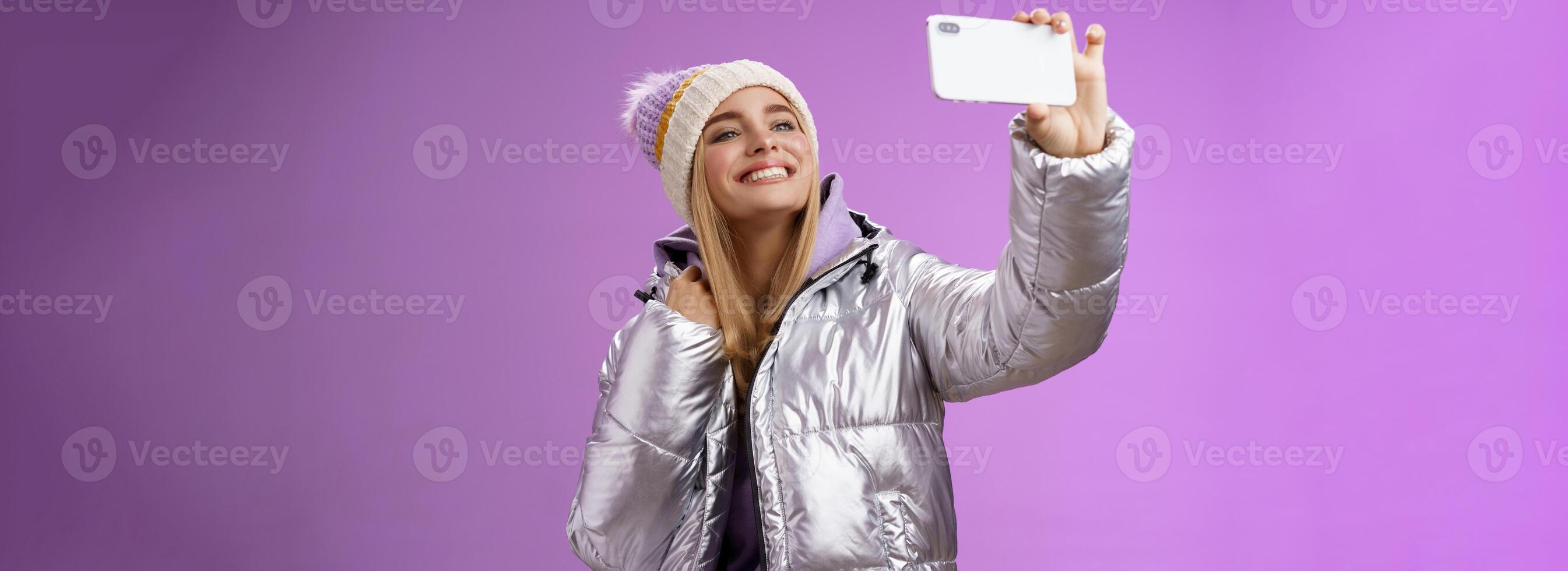Happy delighted carefree fair-haired charming european woman in silver winter jacket hat raising smartphone horizontally taking selfie smiling mobile phone display, purple background photo
