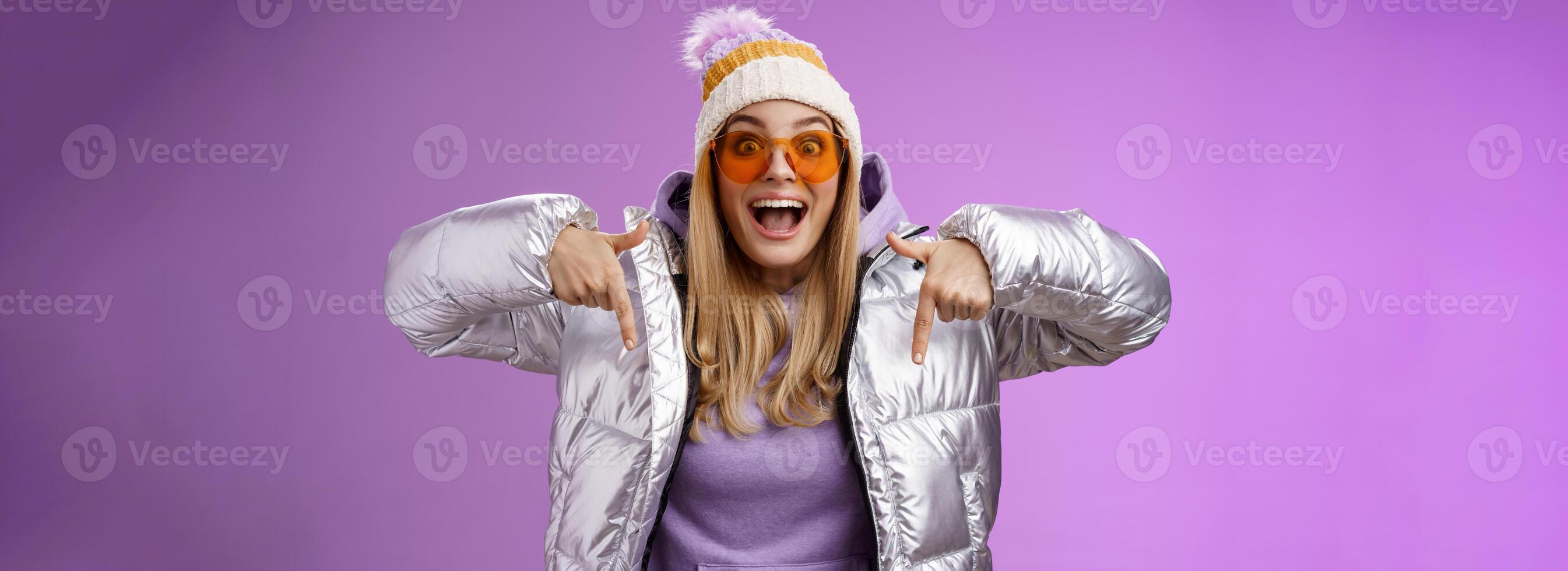 Excited impressed good-looking blond girl drop jaw amused overwhelmed pointing down index fingers checking out awesome promotion standing surprised thrilled wearing silver winter jacket hat photo