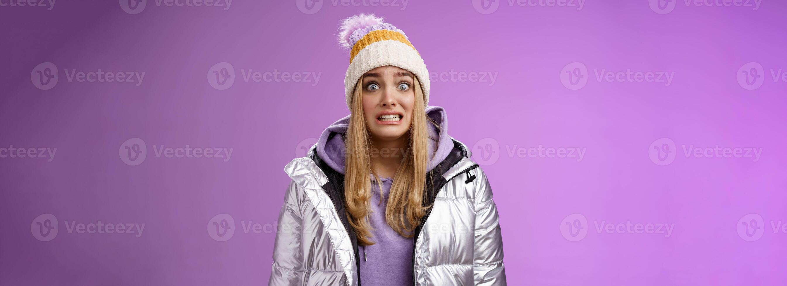 Awkward worried cute timid blond girl in silver jacket hoodie winter hat clench teeth popping eyes camera ooops make mistake standing nervous someone notice, purple background photo