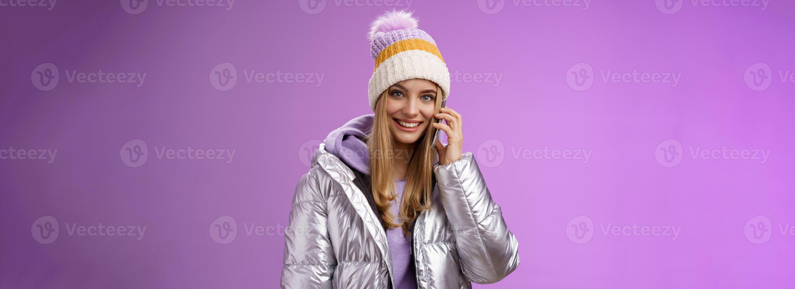 Joyful modern stylish millennial young blond girl talking friends smartphone calling mom from winter resort vacation abroad wearing hat silver jacket hold mobile phone near ear smiling photo