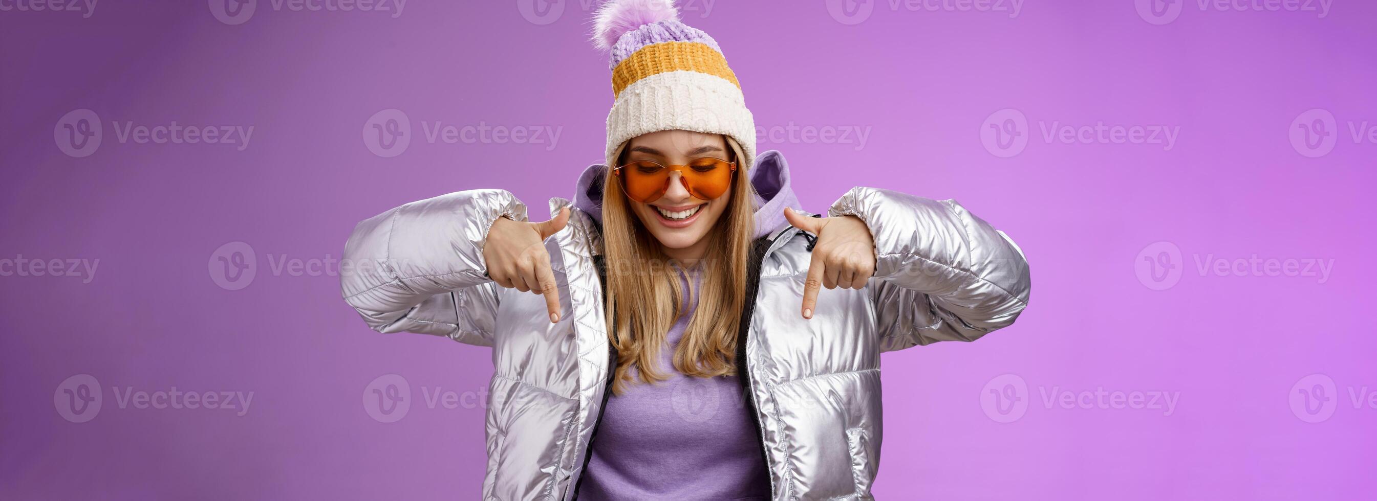 Joyful stylish blond woman in silver jacket stylish sunglasses jat enjoying perfect mountains view snowy vacation resort look pointing down smiling amused having fun feel happy, purple background photo