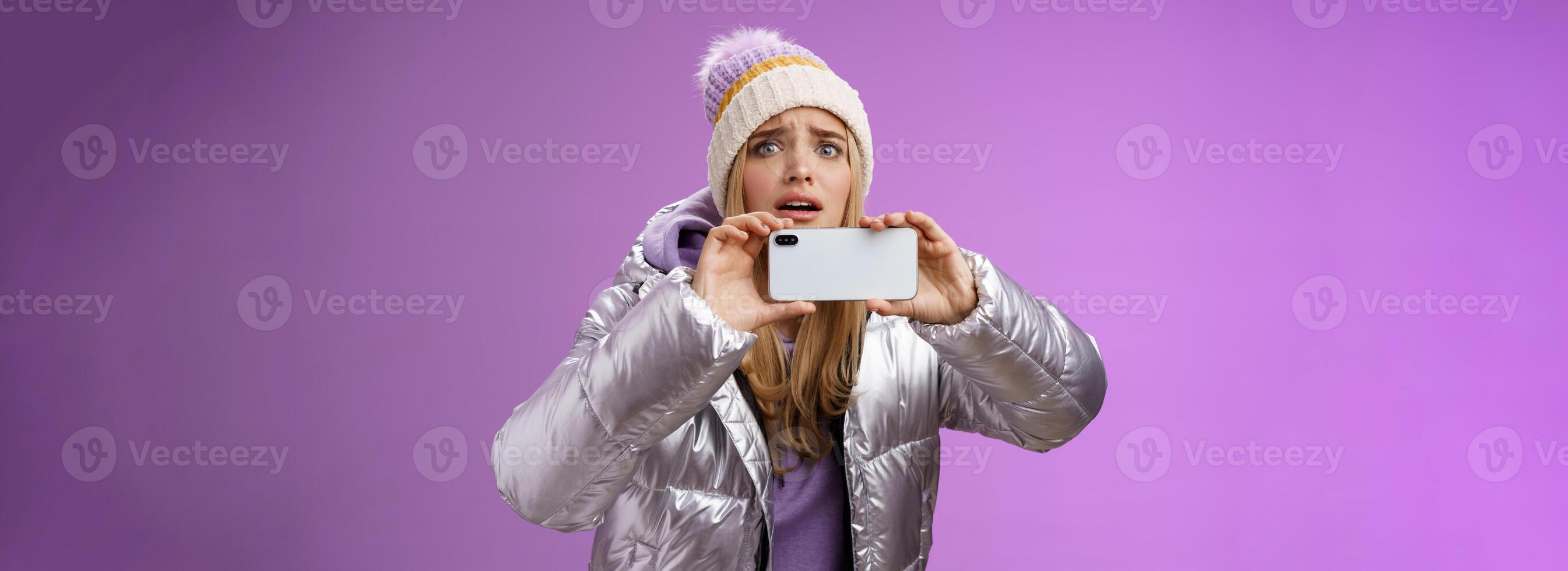 intenso preocupado rubio niña participación móvil teléfono horizontal grabación vídeo capturar momento compartir amigos Internet Blog utilizando teléfono inteligente cámara disparar fotografía en pie grave púrpura antecedentes foto