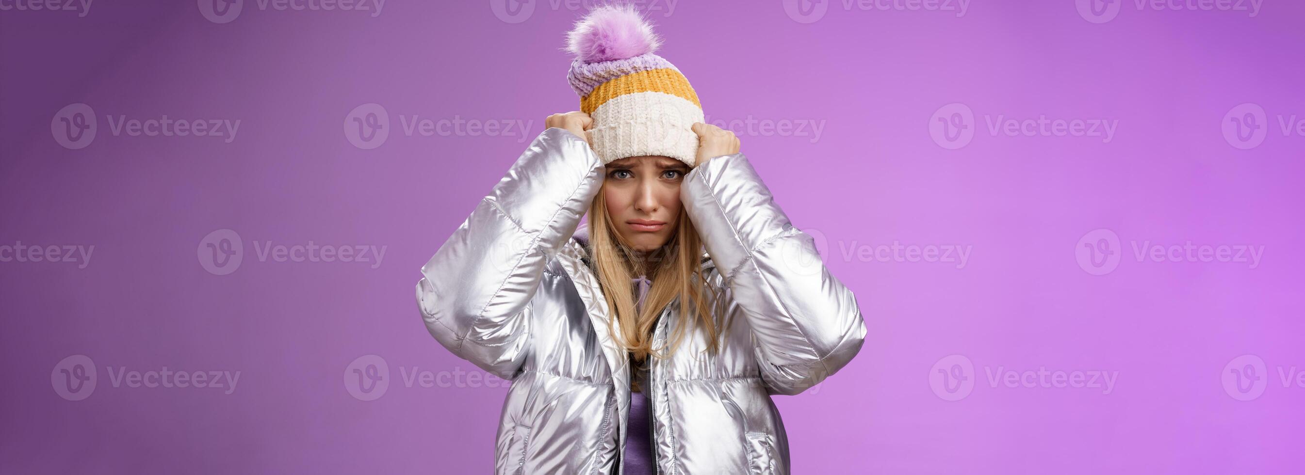 Upset gloomy insulted cute blond girl complaining offensive behaviour pulling hat forehead frowning sulking sad standing miserable pity purple background in warm stylish glittering silver jacket photo