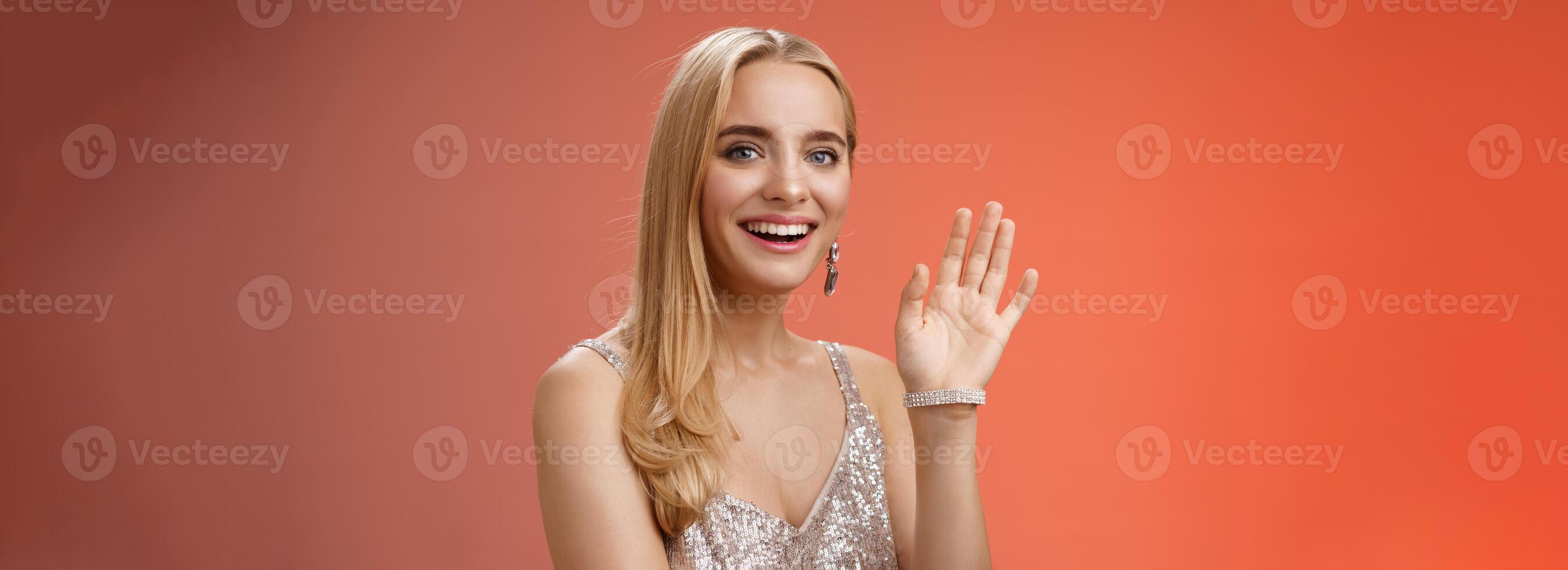 Waist-up studio shot friendly attractive elegant tender blond woman say hi waving raised hand greeting welcoming friend smiling delighted introducing herself hello gesture, red background photo