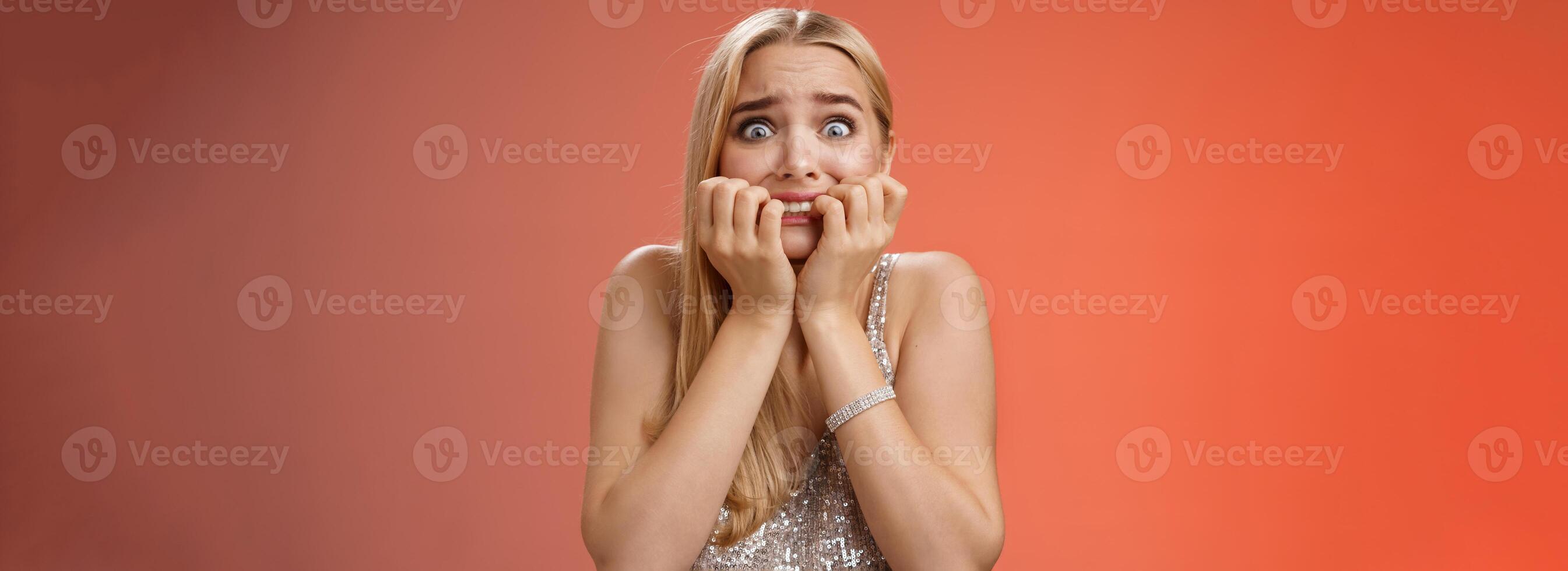 Frightened afraid panicking young cute blond woman biting nails pop eyes scared camera look terrified stunned speechless stare forward standing red background gasping trembling fear photo