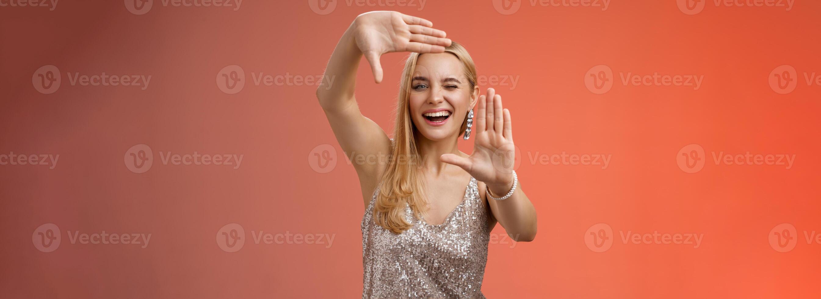 Sassy good-looking creative european blond woman in silver glittering dress winking cheeky smiling confident search inspiration around make frame hand search location take cool shot, red background photo