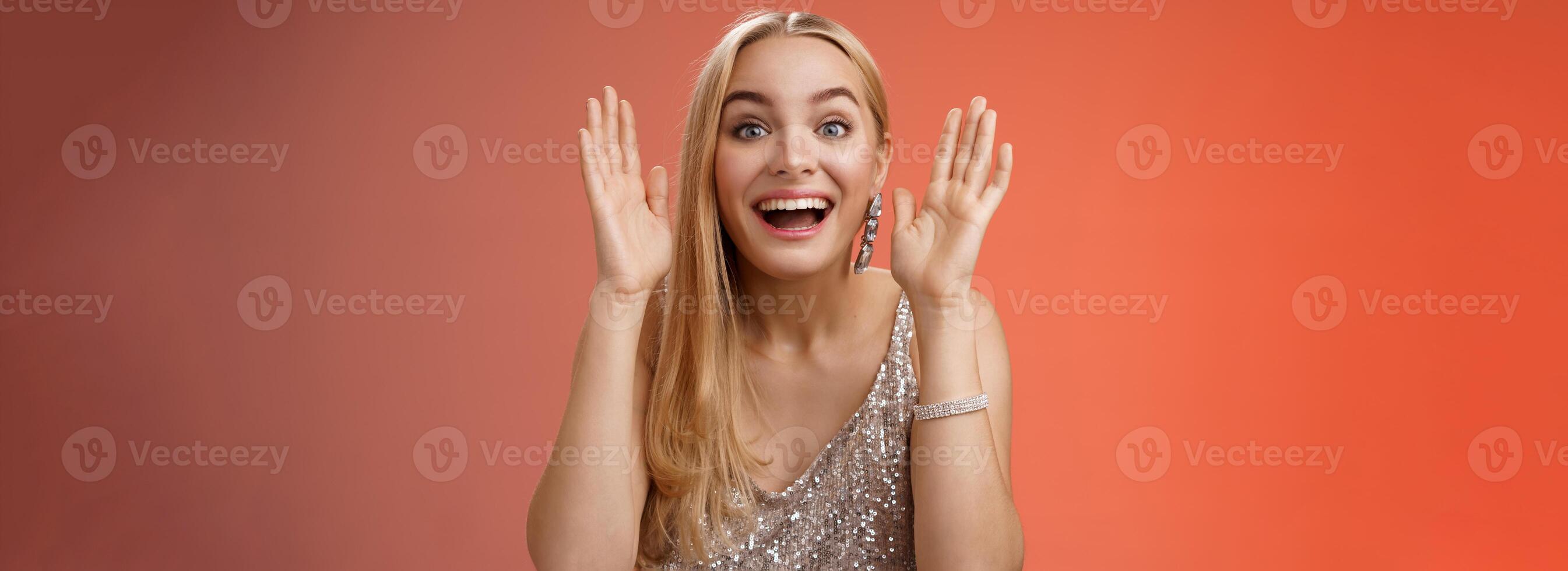 Impressed charming entertained blond european 25s woman in stylish silver glamour dress having fun playing peekaboo smiling joyfully hold palms near face laugh amused, standing red background photo