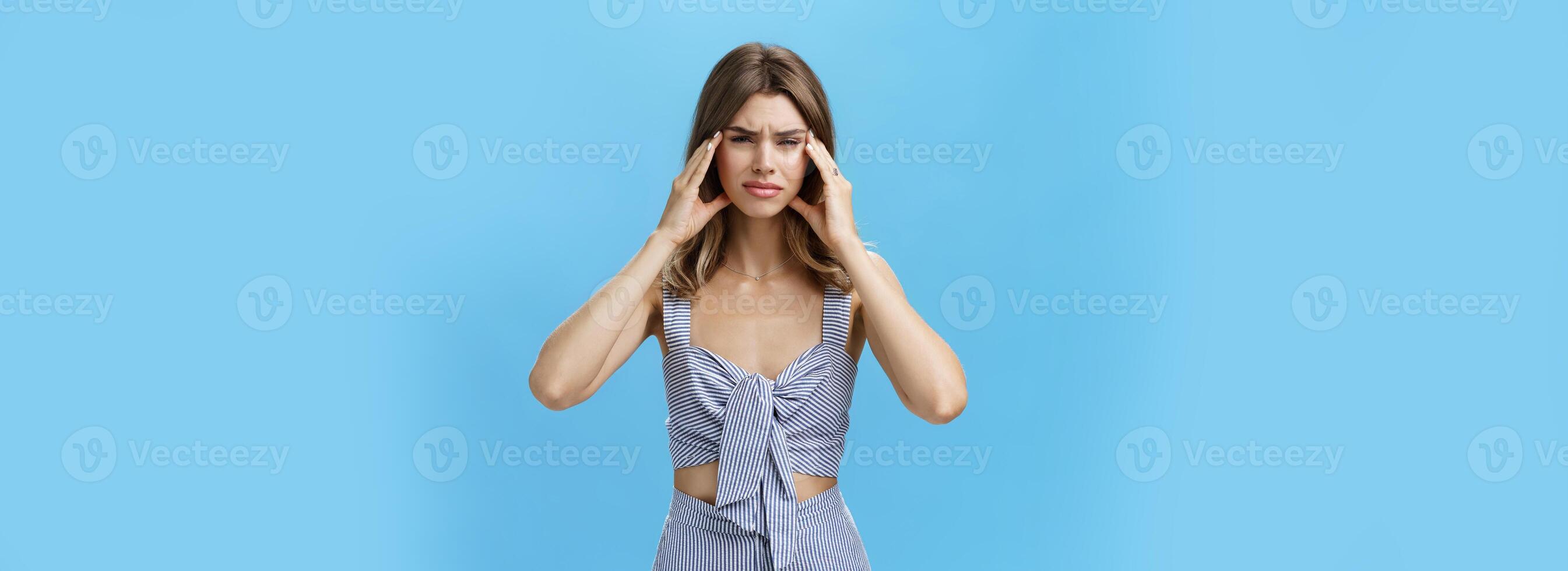 Woman having mental breakdown about to cry having uneasy troublesome situation holding hands on temples frowning making miserable expression suffering from migraine or headache over blue wall photo