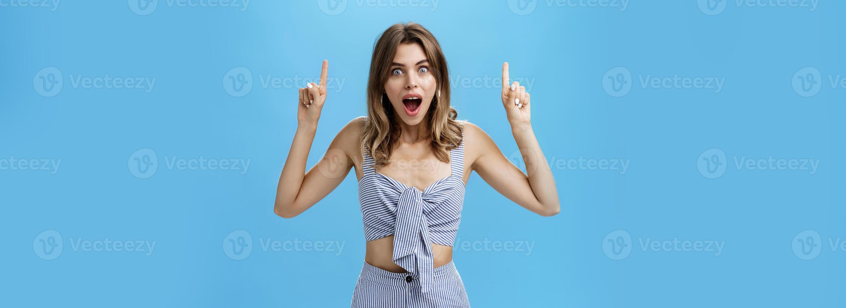 Woman getting surprised dropping jaw from amazement smiling broadly staring at camera astonished raising hands and pointing up at awesome copy space posing against blue background photo