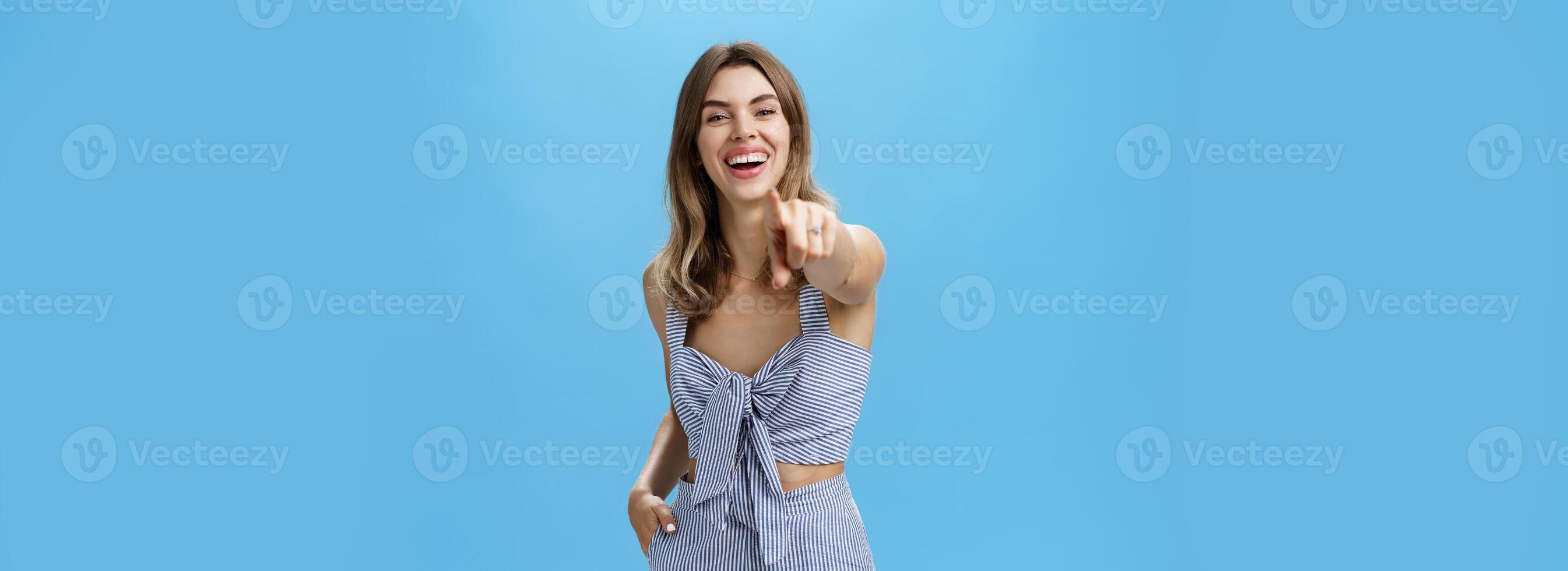 Haha your are funny. Portrait of amused carefree attractive young female in matching outfit pointing at camera as if having fun over photographer laughing out loud being in good mood over blue wall photo