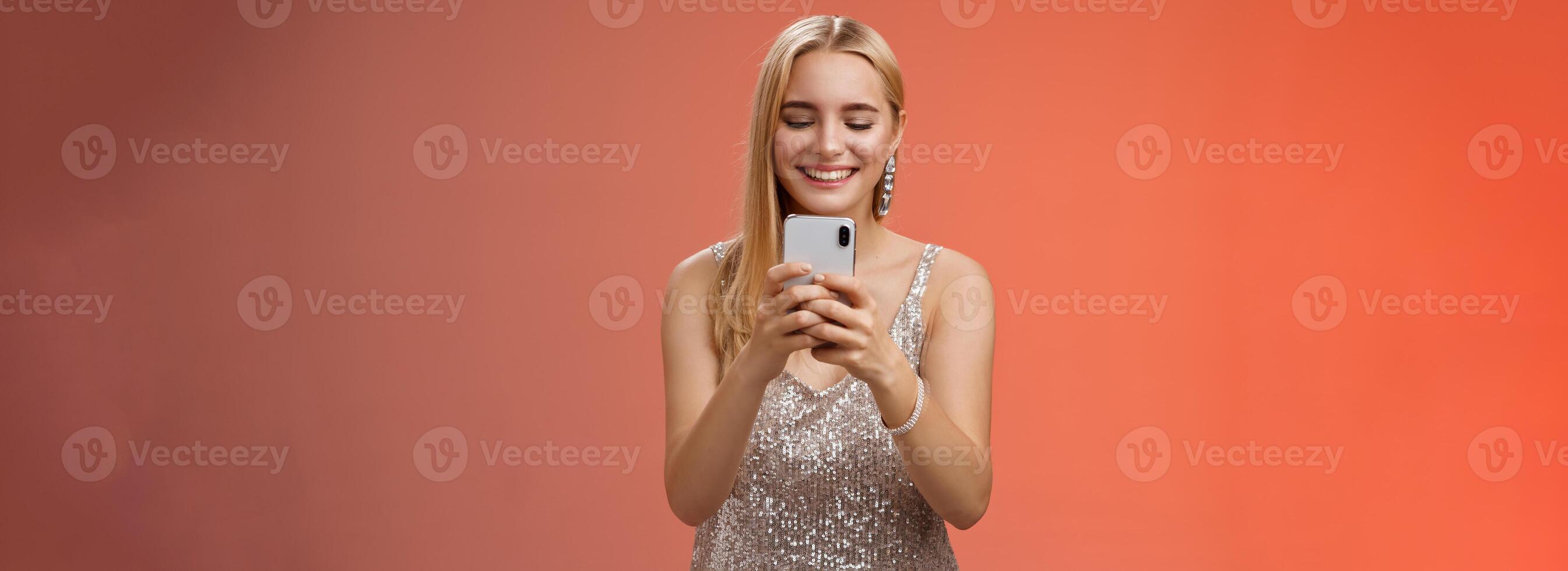 Delighted tender glamour blond woman in silver stylish glittering dress brilliand earrings holding smartphone taking photo friend capture moment celebration nightclub, red background smiling