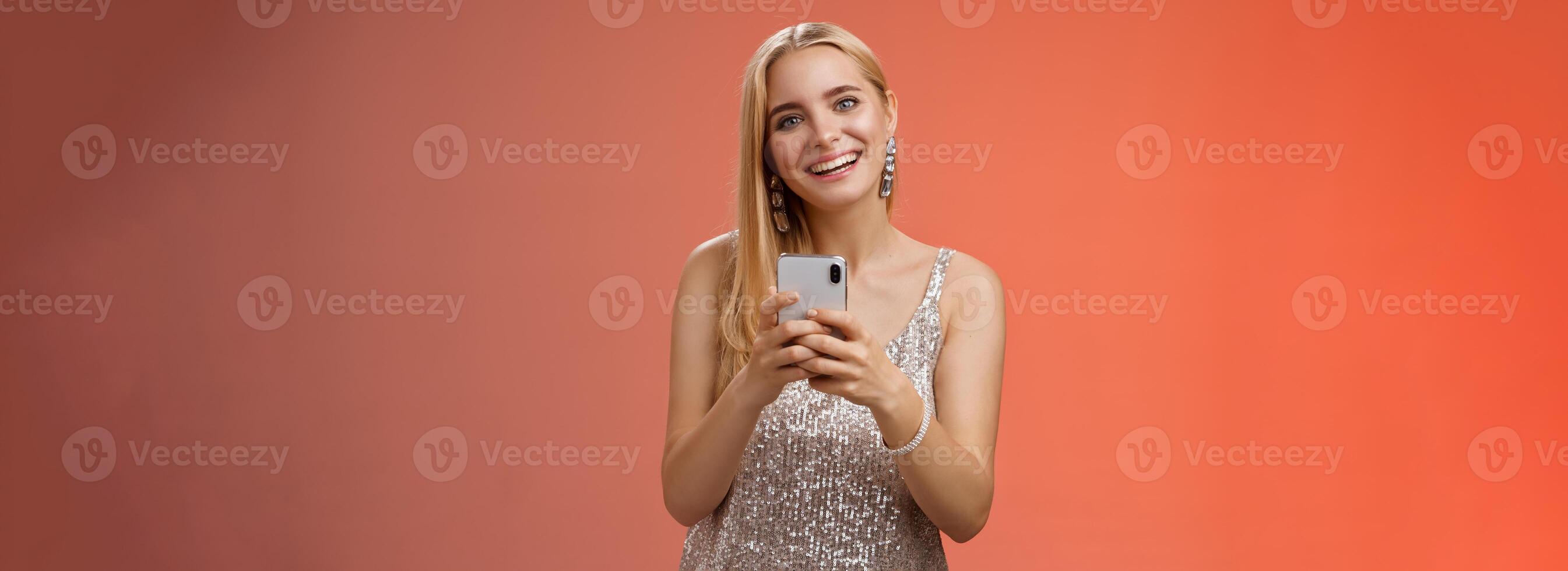 Charming elegant young 25s woman in silver dress brilliant accessorize holding smartphone messaging using app order taxi after nightclub friends party standing carefree smiling camera joyfully photo