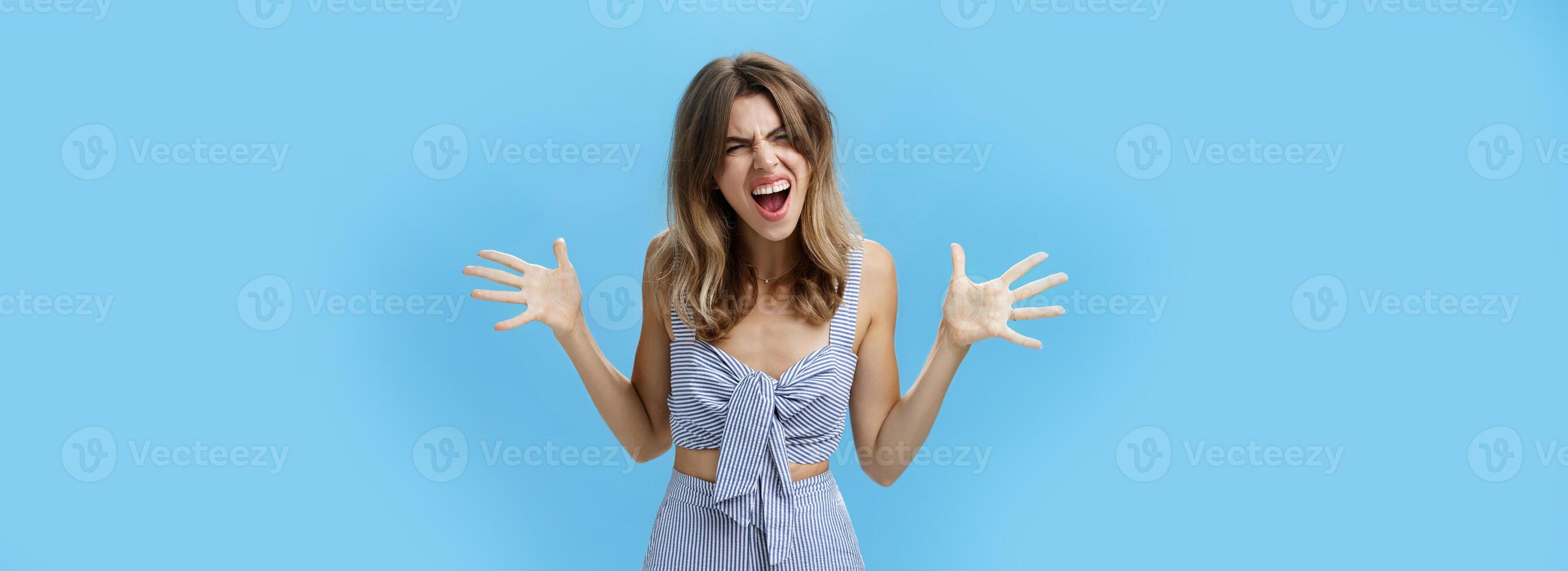 Agressive fed up girlfriend with messy hair and gap teeth yelling shaking raised hands sideways screaming in anger and hate being sick and tired of lies arguing over blue background photo