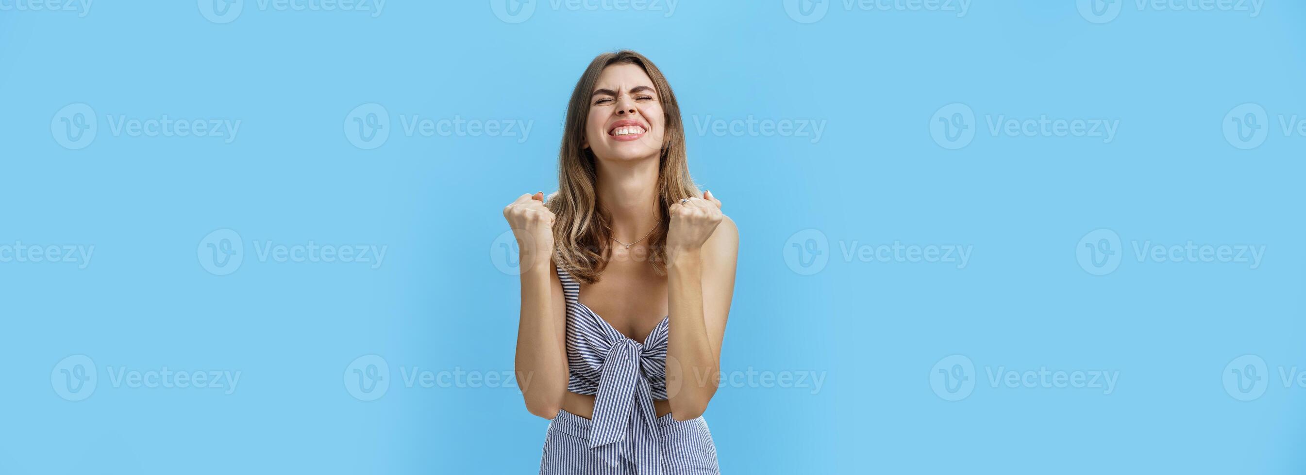 Girl triumphing raising hands in cheer from happiness and victory standing pleased and excited with closed eyes and raised head celebrating successful deal posing delighted against blue background photo