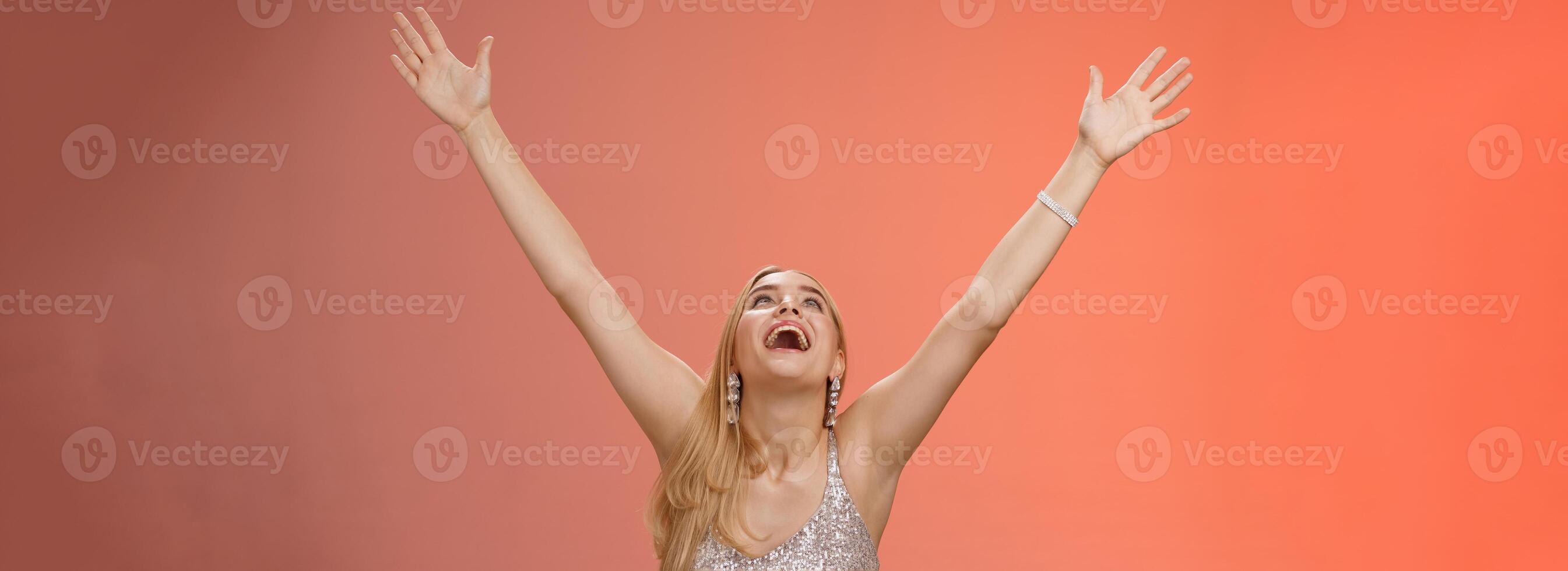 Happy emotive smiling overwhelmed young blond woman in silver dress raise hands sky thank god joyfully signed contract got job rejoicing red background celebrating victory good news, triumphing photo