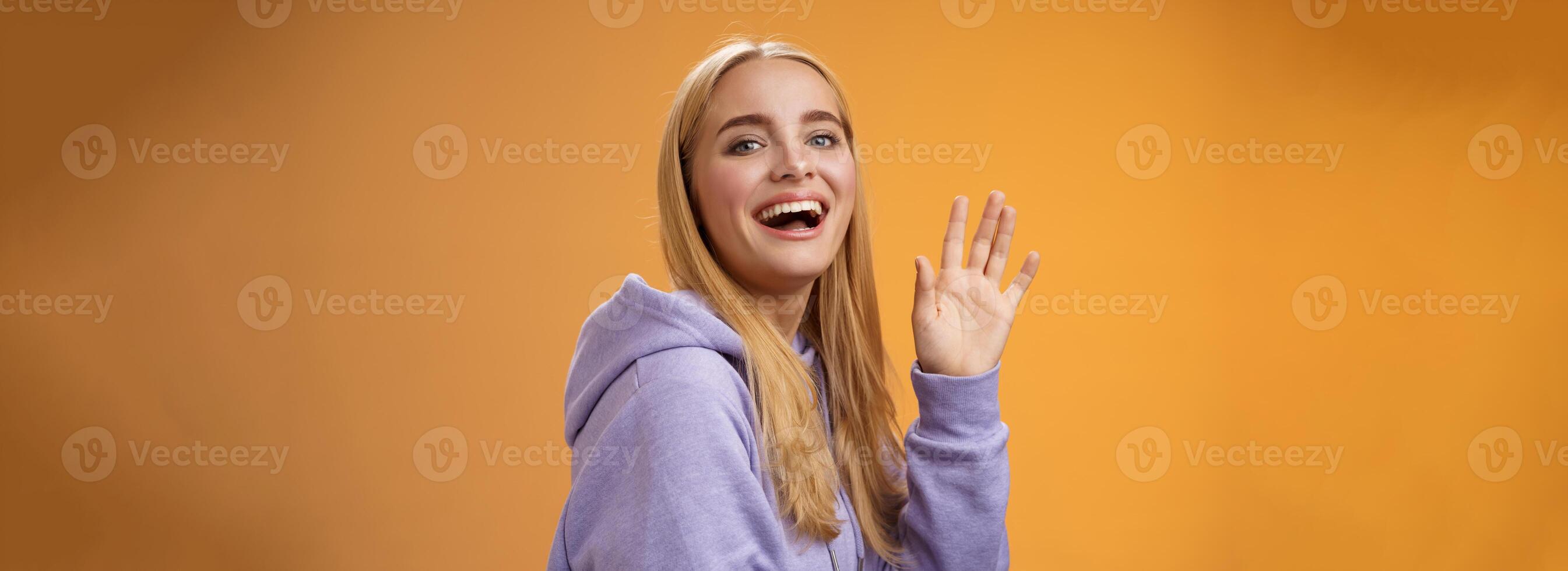 Waist-up friendly lucky cute blond girl waving palm happily smiling turning camera greet friend say hello hi see familiar person welcoming newcomers joyfully, standing orange background photo