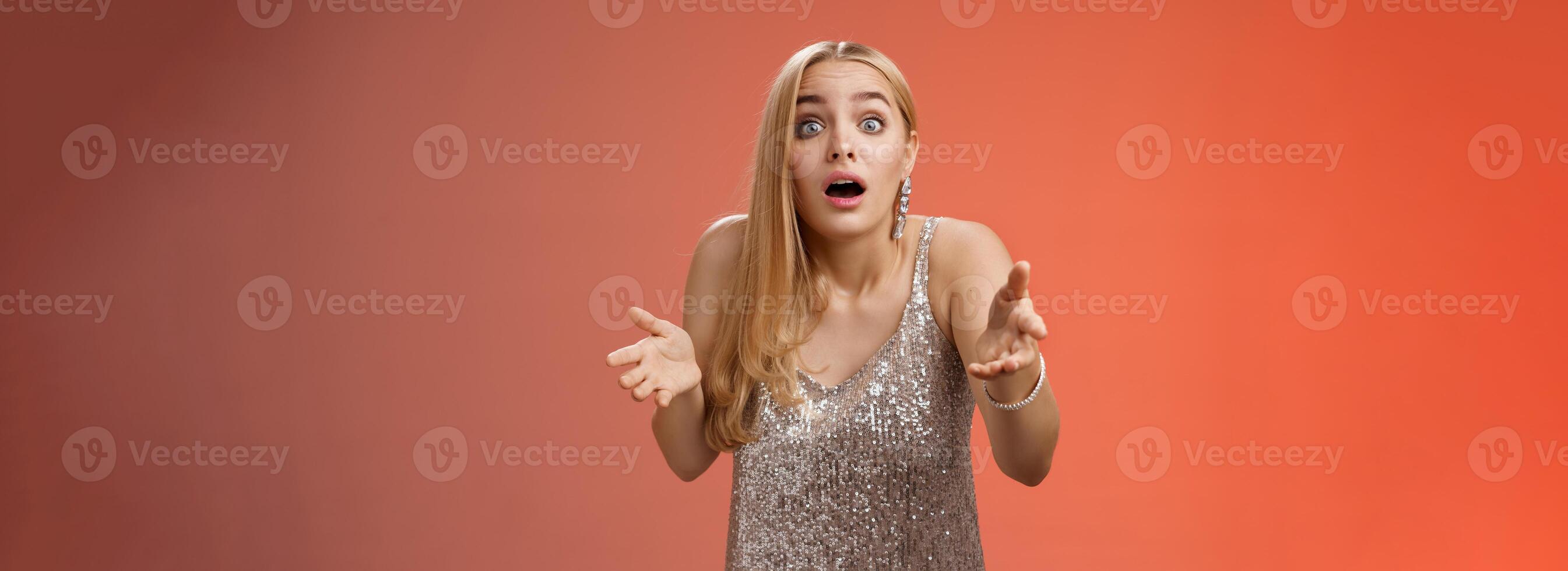 Impressed overwhelmed blond woman retelling exciting story gesturing astonished widen eyes shrugging explaining what happened talking passionately in silver glamour dress standing red background photo