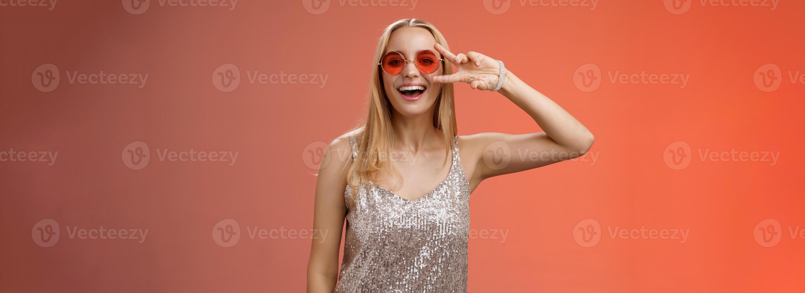 energizado simpático guapo rubio 25 s mujer en plata elegante vestir Gafas de sol espectáculo paz victoria gesto despreocupado sonriente cámara tener divertido disfrutando frio fiesta, en pie rojo antecedentes foto