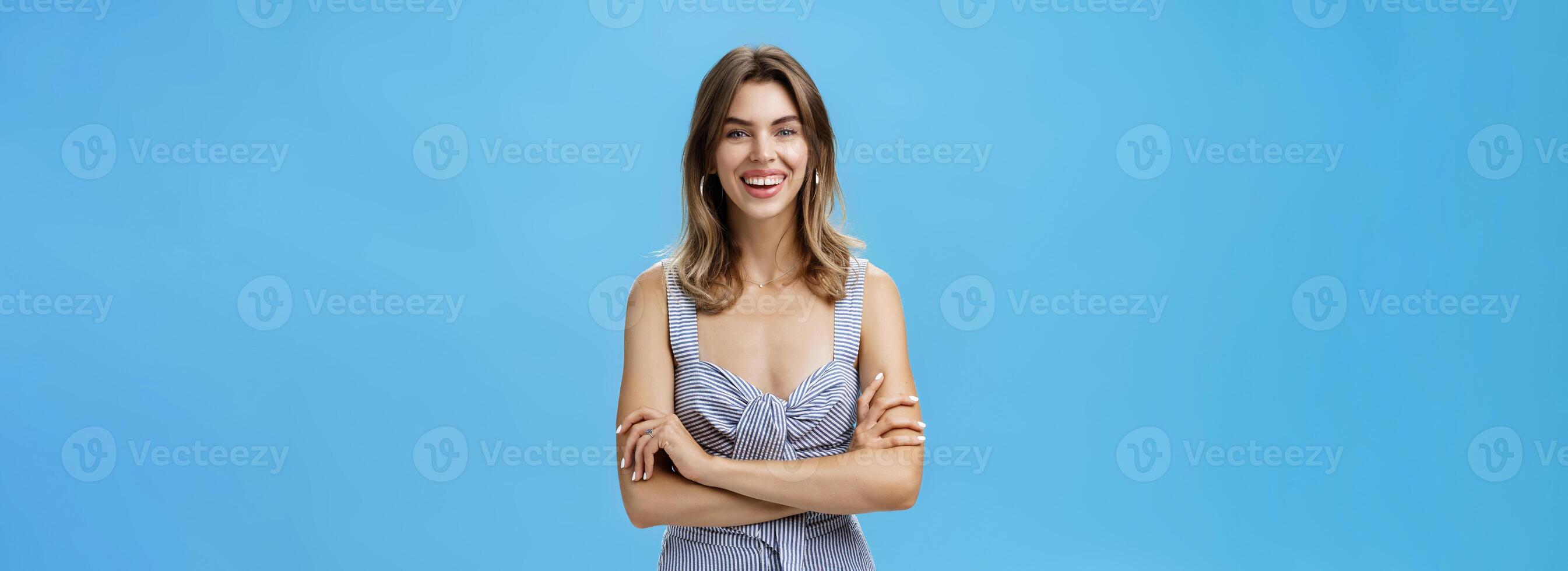Happy charismatic adult european woman with cute gap teeth smiling broadly crossing hands against chest in confident pose having good self-esteem being sure in own natural beauty over blue background photo