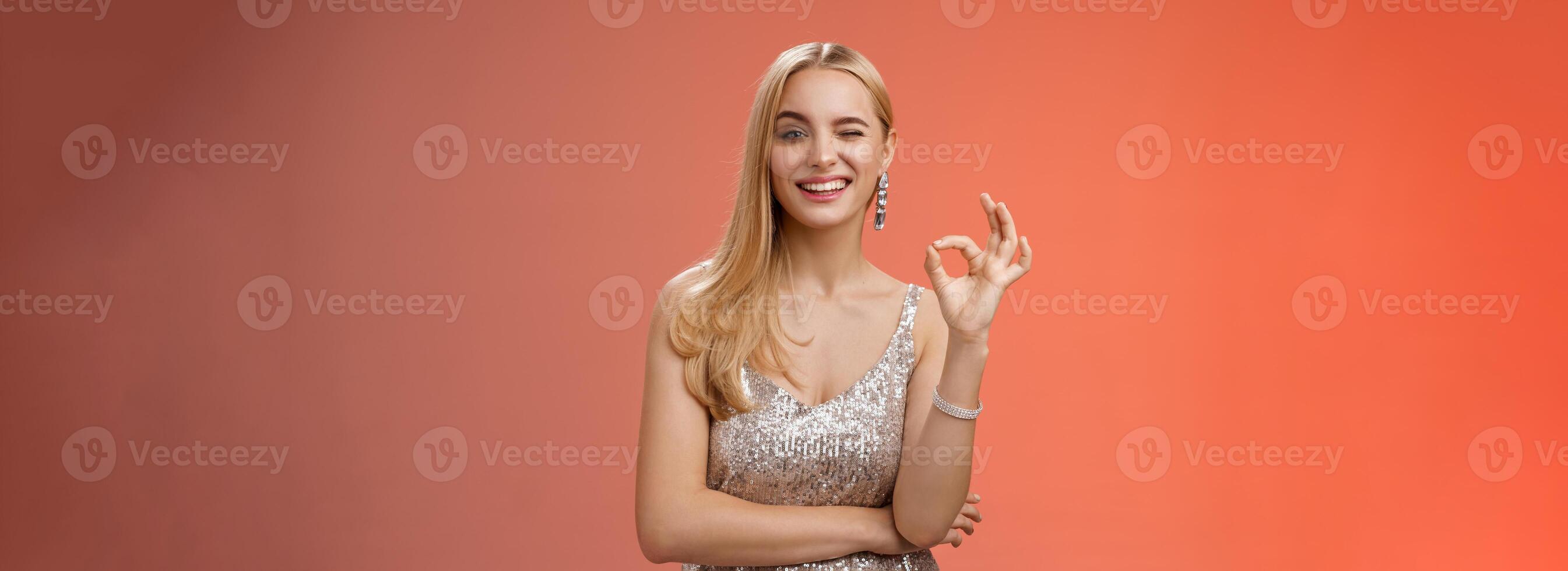 Sassy blond young woman in stylish luxury silver dress winking take things under control show okay no problem gesture make ok sign, like awesome party praise excellent effort, red background photo