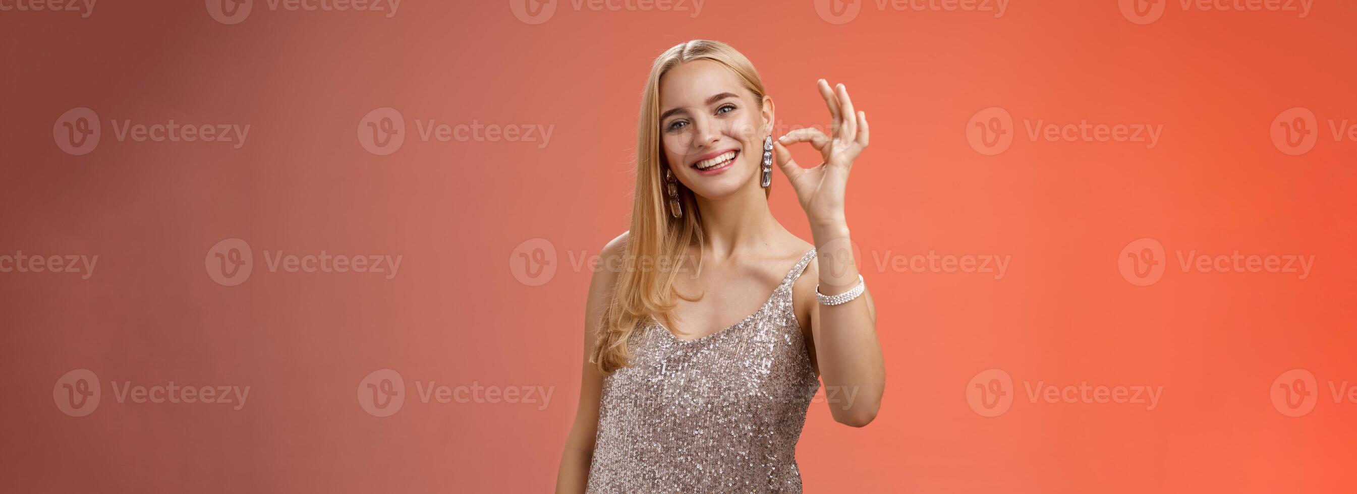 Optimistic attractive stylish blond woman in silver dress show okay ok gesture smiling broadly give approval like recommend cool store buy outfit birthday party, standing red background peleased photo