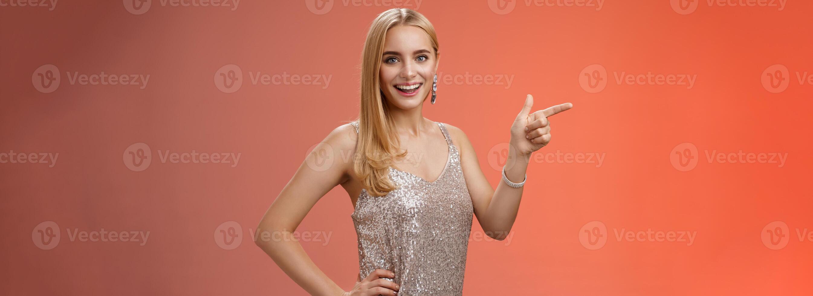 Curious attractive caucasian blond young 25s girl in silver shiny luxurious dress hold hand waist confident pointing left look camera amused joyful entertained party music concert, red background photo
