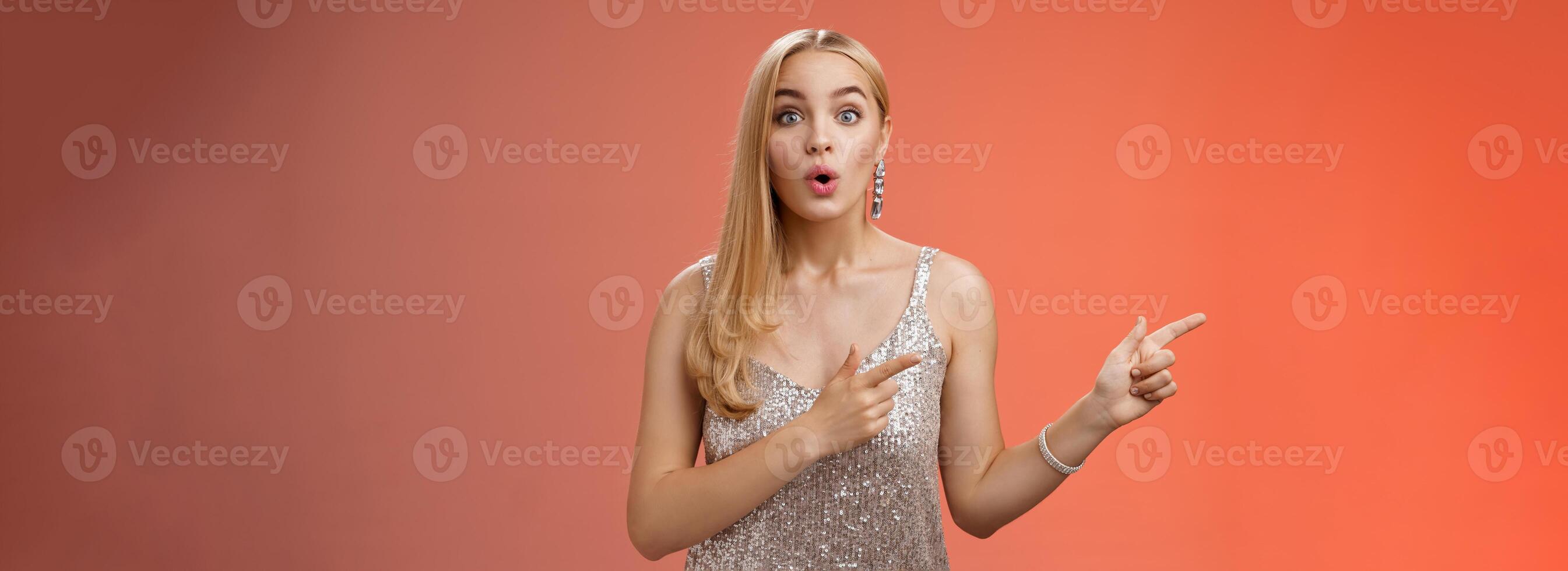 Intrigued excited attractive tender feminine blond european girl in silver stylish dress pointing left amused talking camera widen eyes thrilled asking boyfriend wanna dance shall go check out promo photo