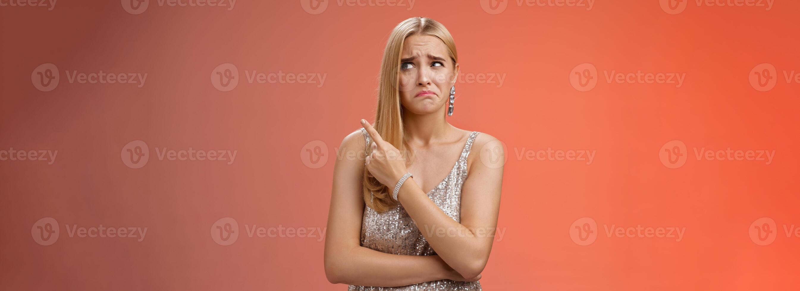 Upset doubtful unsure cute blond funny glamour girl in silver evening dress cringing grimacing hesitant pointing looking upper right corner suspicious, uncertain standing red background photo