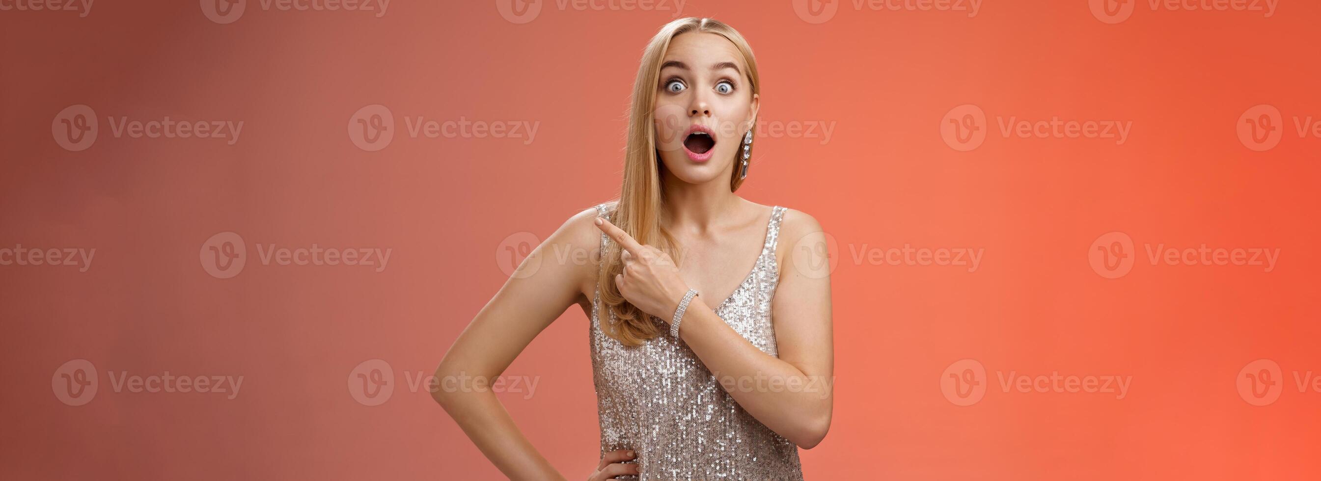 Amused speechless impressed european blong girl drop jaw widen eyes thrilled pointing upper right corner excited amazed standing red background gossiping wealthy bartender wear silver shiny dress photo