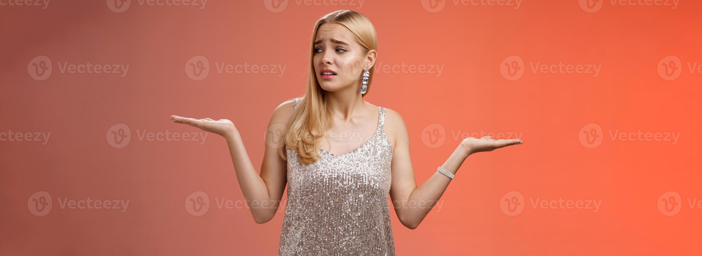 Anxious insecure young cute blond girl feel lost unsure shrugging hands sideways weighing decisions look right seek help standing lost nervous hesitating red background in silver glittering dress photo