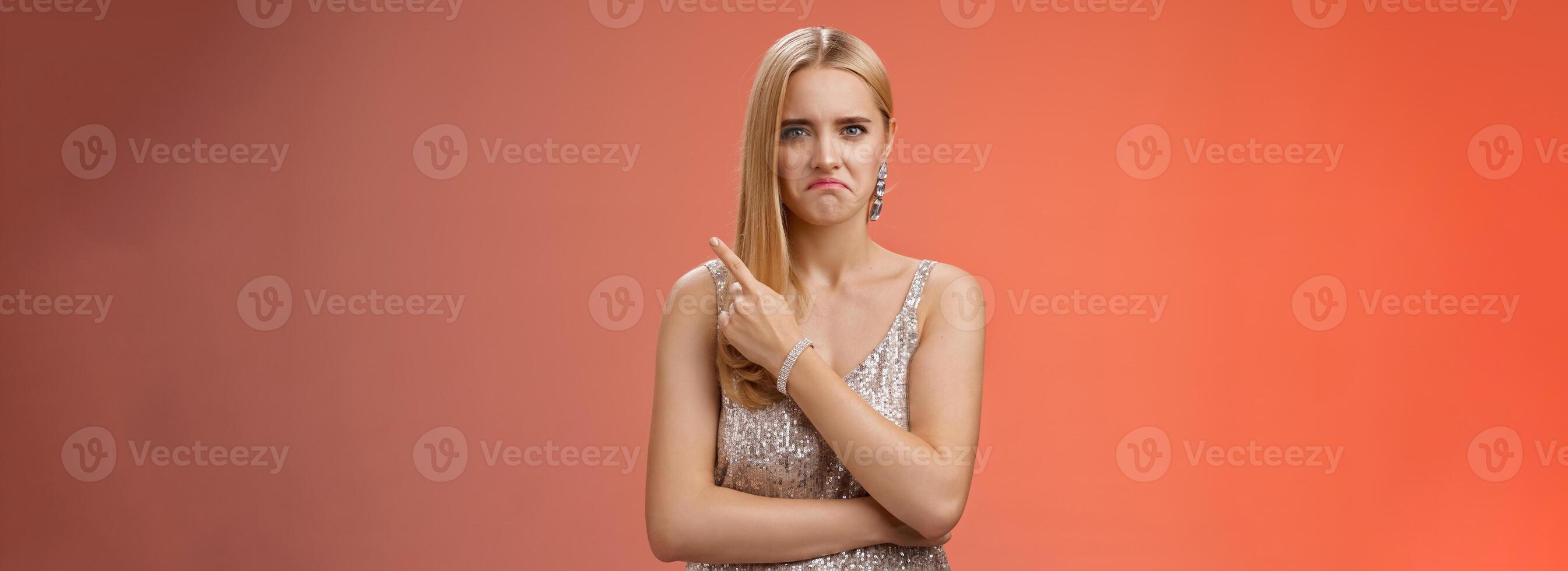 Jealous displeased angry young revengeful blond ex-girlfriend in luxurious silver shiny dress frowning pouting pointing upper right corner displeased pissed standing fed up upset red background photo