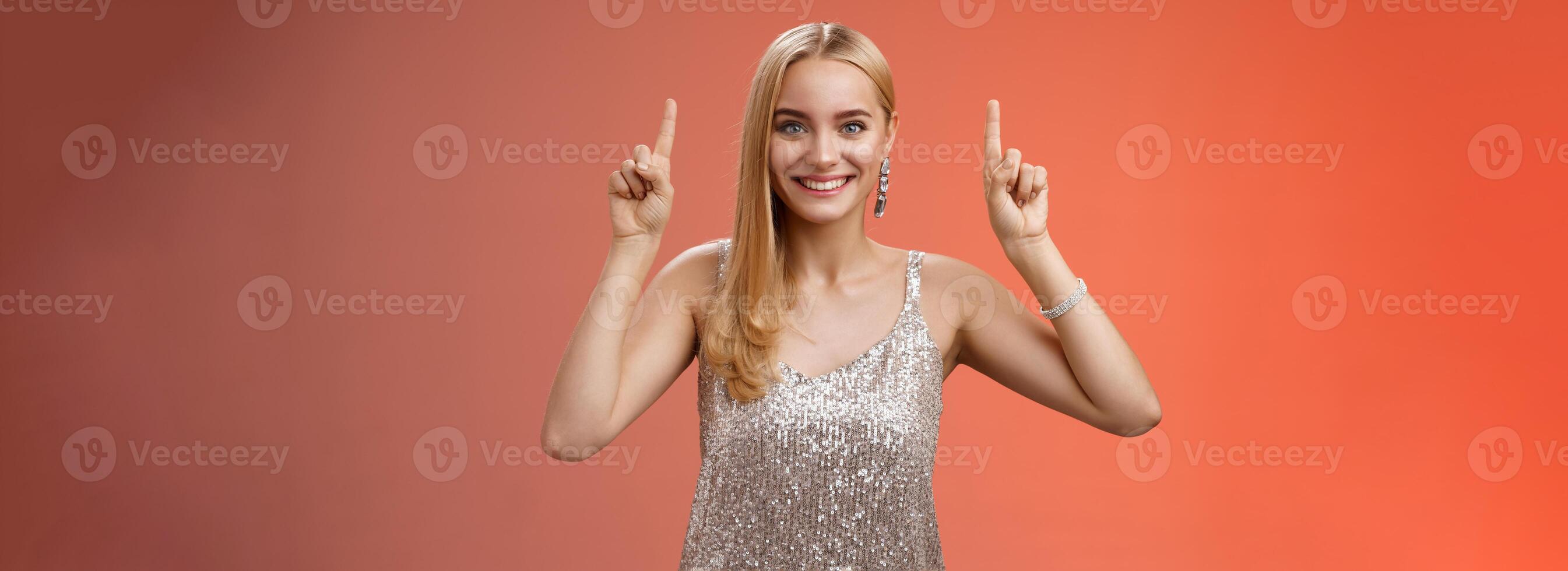 Excited gorgeous blond european woman in silver shiny elegant dress raise hands pointing up showing impressive incredible advertisement smiling happily thrilled wanna take closer look, red background photo