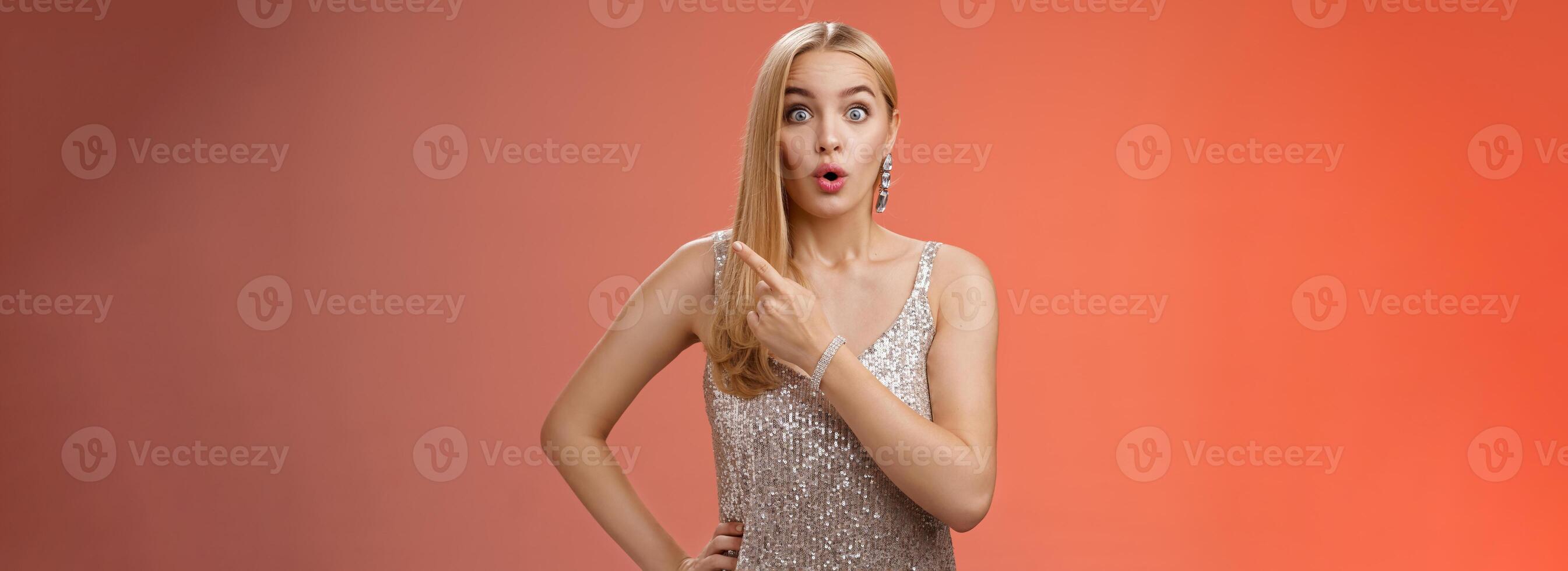 Wondered amused attractive blond woman talking party widen eyes say wow pointing questioned right direction see famous person standing surprised impressed gaze camera, red background photo